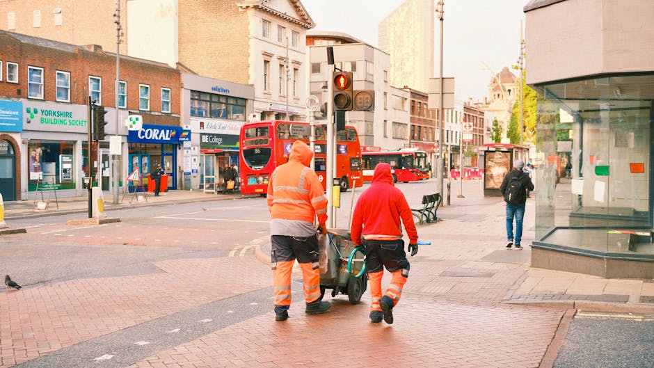 Two street cleaning workers dressed in high-visibility orange and red uniforms are walking along a city sidewalk, pushing a wheeled trash bin. The surrounding environment includes a busy urban landsca