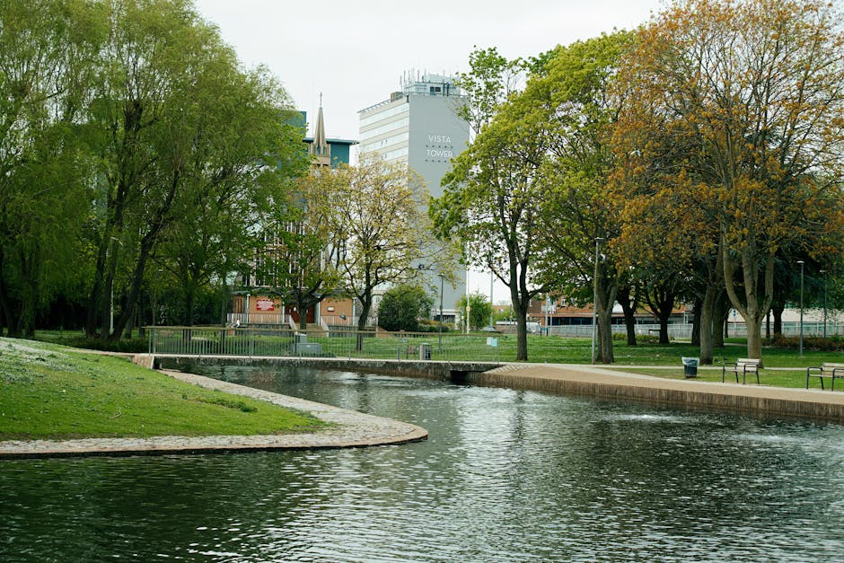 A scenic riverside park in Erith displaying a narrow, gently flowing waterway bordered by a paved walkway and a grassy bank. The park features a variety of deciduous trees, some with fresh green leave