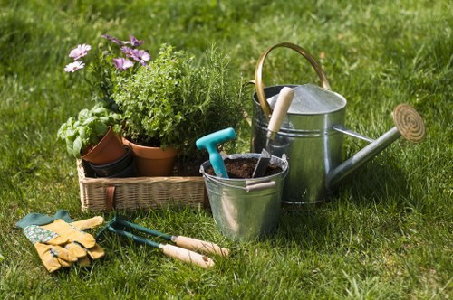 A neatly maintained residential garden in Erith featuring a lush green lawn with even grass density, bordered by a variety of potted plants including purple and green foliage, alongside gardening tools such as trowels and gloves placed on the grass. In the background, there is a wooden garden bed with soil and potted plants, and a metal watering can with a wooden handle positioned nearby, suggesting active gardening or plant care. The environment appears well-kept with natural outdoor lighting, and the overall scene reflects typical garden maintenance activities supported by local gardening services from Garden Maintenance Erith to keep outdoor spaces healthy and attractive.